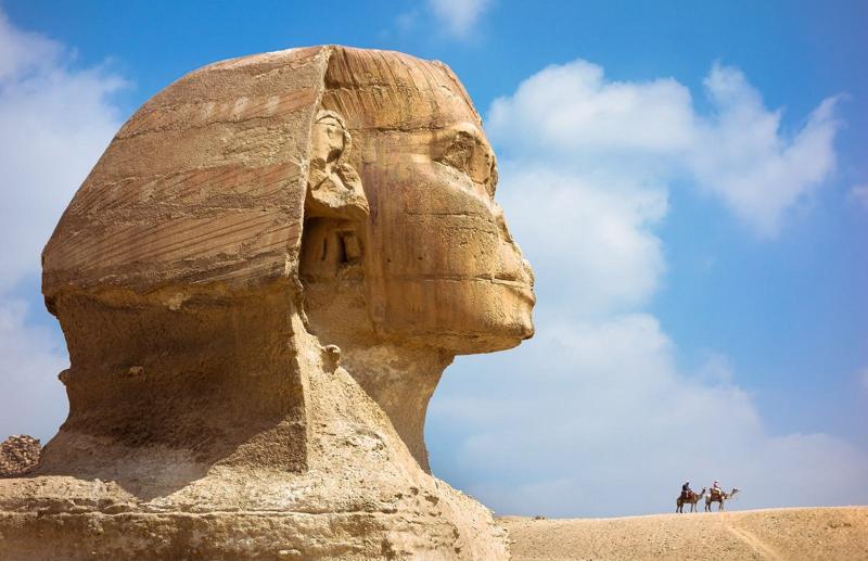 The Great Sphinx of Giza in profile, with camel riders on the desert behind